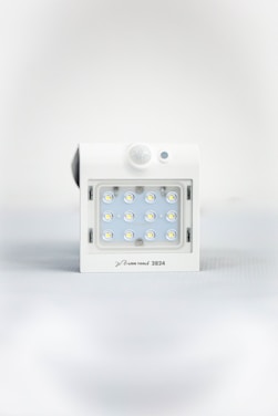 A square white LED light fixture featuring twelve small illuminated LED bulbs arranged in a grid. The fixture has a white casing with a small, round motion sensor above the LED grid and a branding label at the bottom.