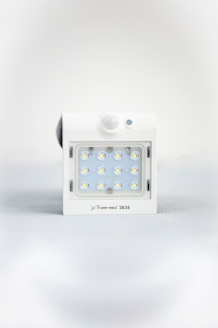 A square white LED light fixture featuring twelve small illuminated LED bulbs arranged in a grid. The fixture has a white casing with a small, round motion sensor above the LED grid and a branding label at the bottom.
