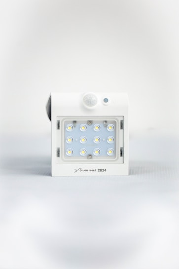 A square white LED light fixture featuring twelve small illuminated LED bulbs arranged in a grid. The fixture has a white casing with a small, round motion sensor above the LED grid and a branding label at the bottom.