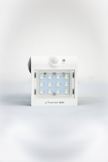 A square white LED light fixture featuring twelve small illuminated LED bulbs arranged in a grid. The fixture has a white casing with a small, round motion sensor above the LED grid and a branding label at the bottom.