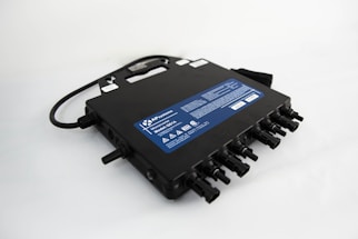 A black electronic device with multiple input and output connections, featuring a handle on top and a blue label displaying technical specifications and safety warnings.