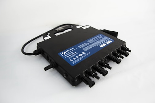 A black electronic device with multiple input and output connections, featuring a handle on top and a blue label displaying technical specifications and safety warnings.