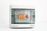 A gray electrical control panel with a transparent cover, inside which are orange and white labeled controls and a prominent red rotary switch.