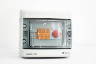 A gray electrical control panel with a transparent cover, inside which are orange and white labeled controls and a prominent red rotary switch.