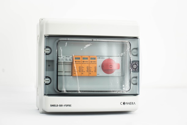 A gray electrical control panel with a transparent cover, inside which are orange and white labeled controls and a prominent red rotary switch.