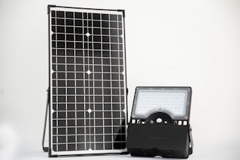 A solar panel stands upright next to a LED floodlight on a simple, clean background. The solar panel features a grid of photovoltaic cells, while the LED light consists of multiple small bulbs within a rectangular frame.