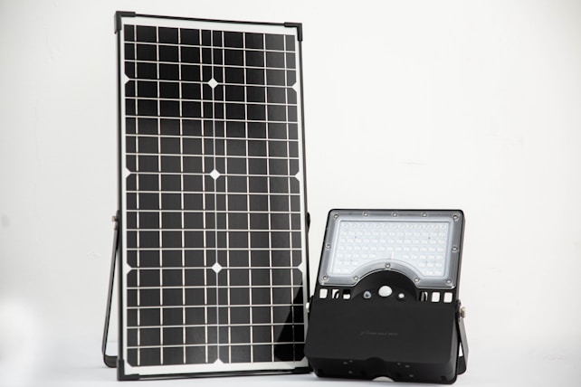 A solar panel stands upright next to a LED floodlight on a simple, clean background. The solar panel features a grid of photovoltaic cells, while the LED light consists of multiple small bulbs within a rectangular frame.