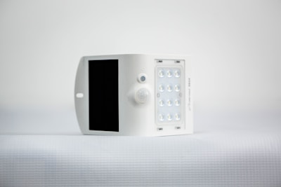 A small, white, rectangular device featuring a solar panel on one side and an array of LED lights on the opposite side. It is positioned against a minimalistic, light background.