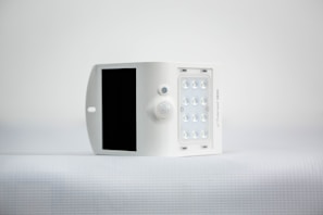 A small, white, rectangular device featuring a solar panel on one side and an array of LED lights on the opposite side. It is positioned against a minimalistic, light background.