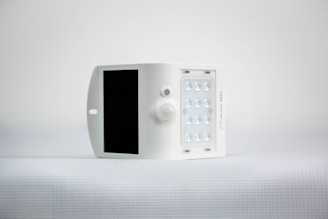 A small, white, rectangular device featuring a solar panel on one side and an array of LED lights on the opposite side. It is positioned against a minimalistic, light background.