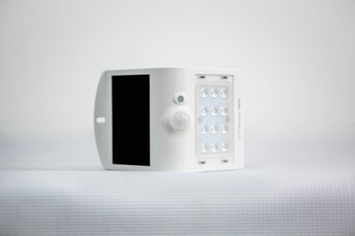 A small, white, rectangular device featuring a solar panel on one side and an array of LED lights on the opposite side. It is positioned against a minimalistic, light background.