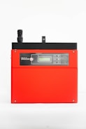A red electronic device with a black top section featuring a small antenna and digital display. The front has a control panel with several buttons and icons, possibly indicating connectivity or settings.