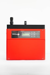 A red electronic device with a black top section featuring a small antenna and digital display. The front has a control panel with several buttons and icons, possibly indicating connectivity or settings.