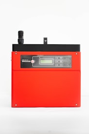 A red electronic device with a black top section featuring a small antenna and digital display. The front has a control panel with several buttons and icons, possibly indicating connectivity or settings.