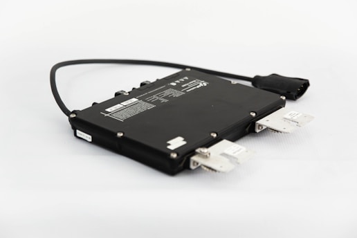 A black electronic device with a rectangular shape, multiple connectors and labels, positioned on a light surface. A cable extends from the side, ending in a plug.