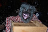 A figure resembling a werewolf with a gray furry appearance and large sharp teeth is perched on a wooden crate. The creature is adorned in a red plaid shirt. In the background, a skull is partially visible, contributing to a spooky atmosphere with dark surroundings.
