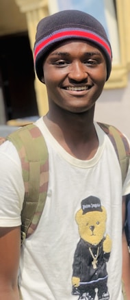 A person wearing a beanie with red and blue stripes, a white T-shirt featuring a bear in a black outfit, and a camouflage-patterned backpack. The person has a cheerful expression.