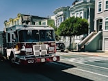 A fire truck labeled S.F.F.D is parked on a residential street with classic Victorian-style homes in the background. The street is sunny and lined with trees. Sidewalks are visible alongside the parked vehicles.