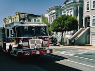 A fire truck labeled S.F.F.D is parked on a residential street with classic Victorian-style homes in the background. The street is sunny and lined with trees. Sidewalks are visible alongside the parked vehicles.
