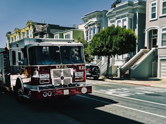 A fire truck labeled S.F.F.D is parked on a residential street with classic Victorian-style homes in the background. The street is sunny and lined with trees. Sidewalks are visible alongside the parked vehicles.