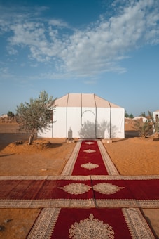 A large white tent is set up in a sandy desert environment, framed by a clear blue sky with scattered clouds. In front of the tent, a long red carpet with intricate patterns stretches across the sand. Two decorative lanterns are placed at the entrance of the tent, while small trees and bushes are visible on either side.