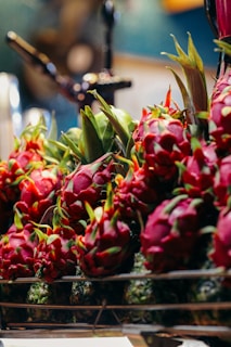 A vibrant display of dragon fruits arranged on a rack. The fruits are bright reddish-pink with green scales, surrounded by a blurred background, suggesting a market or grocery setting. The lighting highlights the fresh and exotic appearance of the dragon fruits.