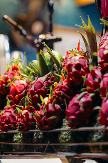 A vibrant display of dragon fruits arranged on a rack. The fruits are bright reddish-pink with green scales, surrounded by a blurred background, suggesting a market or grocery setting. The lighting highlights the fresh and exotic appearance of the dragon fruits.