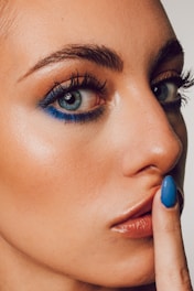 A close-up of a person's face focusing on bright blue eye makeup and well-groomed eyebrows. The person's skin has a smooth texture, and they have glossy lips. A finger with blue nail polish is placed in front of the lips in a gesture that suggests silence.