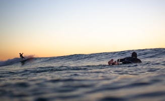 A surfer glides smoothly along a breaking wave, silhouetted against a vibrant horizon during sunset. Nearer in the foreground, another individual lies on a surfboard, watching the surfer in action. The water appears calm except for the gentle swell, and the sky is a gradient of warm orange fading into blue.