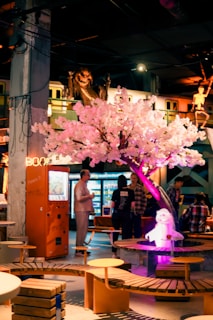 A vibrant, indoor scene featuring a striking pink cherry blossom tree with illuminated lights. A group of people is gathered around, engaged in conversation. The setting includes modern, eclectic furniture and decor elements, such as an orange vending machine and seating made from wood and metal. A playful, lit-up dog sculpture adds to the whimsical atmosphere. In the background, hints of an animated setting with a creature or monster and a skeleton create a unique ambiance.