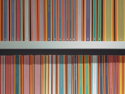 A neatly organized bookshelf displays a collection of brightly colored book spines, each predominantly featuring bold stripes with titles printed along the vertical edge. The books are primarily classic literature from a well-known publisher, with repeating small logo icons visible at the base of each spine.