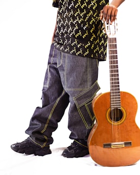 A person wearing baggy jeans and a patterned shirt stands beside a classical guitar. The guitar is leaning against their leg, and the person is holding its neck. Black sneakers complement the casual and laid-back attire.