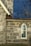 A detailed section of a stone building wall with a pointed arch stained glass window. The wall features large grey stones with mortar, and the window has decorative blue patterns on the glass. The roof is covered with dark shingles and features a beige gutter downspout.