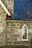 A detailed section of a stone building wall with a pointed arch stained glass window. The wall features large grey stones with mortar, and the window has decorative blue patterns on the glass. The roof is covered with dark shingles and features a beige gutter downspout.