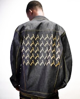 A person is wearing a denim jacket adorned with a pattern of yellow and white cursive letters. The individual is photographed from behind, highlighting the jacket's design against a plain white background.