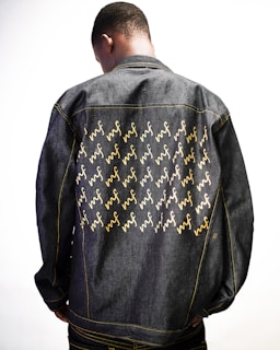 A person is wearing a denim jacket adorned with a pattern of yellow and white cursive letters. The individual is photographed from behind, highlighting the jacket's design against a plain white background.