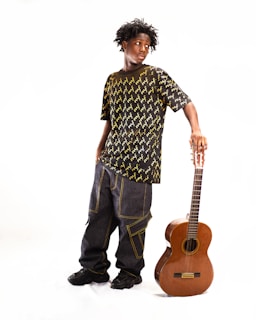 A young person stands in a relaxed pose, wearing a black shirt with a yellow pattern and baggy jeans. They are leaning on a classical guitar which rests vertically on the floor. The background is plain and bright, highlighting the subject.