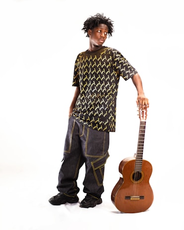 A young person stands in a relaxed pose, wearing a black shirt with a yellow pattern and baggy jeans. They are leaning on a classical guitar which rests vertically on the floor. The background is plain and bright, highlighting the subject.