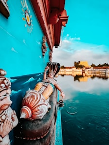 A vibrant and detailed mural featuring a reclining deity with intricate ornamentation, set against a turquoise wall. The scene is by a calm body of water reflecting a clear sky and distant architecture, including a temple structure.
