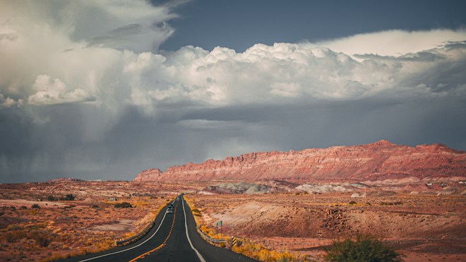 An open highway stretches into the distance, flanked by expansive desert landscapes with reddish cliffs under a dramatic sky filled with thick, gray clouds.