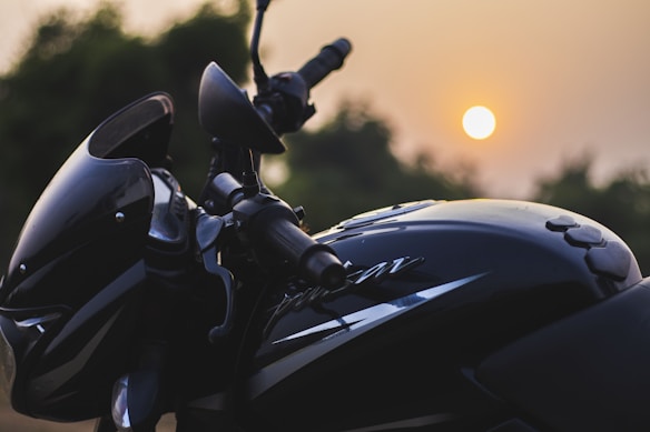 The image features a close-up view of a motorcycle, highlighting its sleek design and glossy black finish. The sun is setting in the background, casting a warm glow and creating a serene atmosphere. The motorcycle's handlebars and rearview mirrors are visible, with trees blurred in the background.