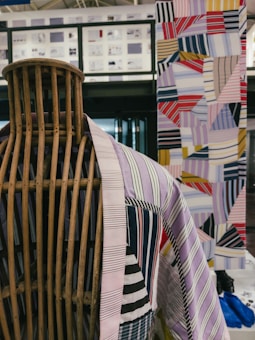 A wooden mannequin is adorned with a colorful, striped textile garment. In the background, a quilt or patchwork design is displayed featuring an array of geometric patterns and vibrant colors. The setting appears to be a contemporary interior space with modern architectural elements.
