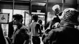 A black and white photo capturing a dynamic street scene at night. Several people are moving in and out of a glass door, which displays the number 107 above it. A photographer in the foreground is taking a picture, holding a camera with a large flash. Posters and reflections are visible in the windows, adding to the urban ambiance.
