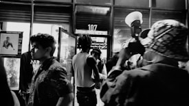A black and white photo capturing a dynamic street scene at night. Several people are moving in and out of a glass door, which displays the number 107 above it. A photographer in the foreground is taking a picture, holding a camera with a large flash. Posters and reflections are visible in the windows, adding to the urban ambiance.