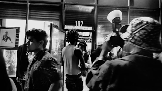 A black and white photo capturing a dynamic street scene at night. Several people are moving in and out of a glass door, which displays the number 107 above it. A photographer in the foreground is taking a picture, holding a camera with a large flash. Posters and reflections are visible in the windows, adding to the urban ambiance.