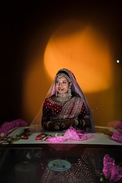 A bride in traditional attire adorned with intricate jewelry stands in a well-lit environment. The focus is on her smiling face framed by a veil, with rich red and gold embroidered fabric creating an elegant and festive appearance. The background features a warm, glowing circular light, enhancing the celebratory atmosphere.