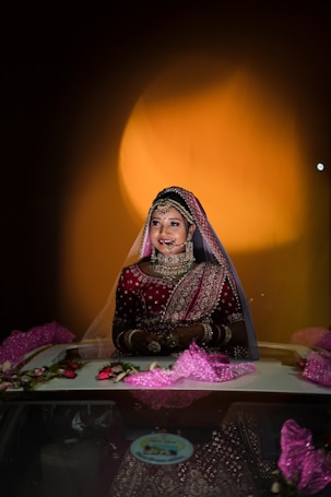 A bride in traditional attire adorned with intricate jewelry stands in a well-lit environment. The focus is on her smiling face framed by a veil, with rich red and gold embroidered fabric creating an elegant and festive appearance. The background features a warm, glowing circular light, enhancing the celebratory atmosphere.