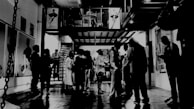 An art gallery setting with people gathered in a dimly lit, industrial-style space. The floor is reflective, and there are artworks displayed on the walls. Some people are standing in groups, possibly engaging in conversation or examining the art pieces. A mezzanine level with additional figures is visible, adding depth to the scene. The overall atmosphere is casual yet sophisticated.