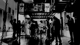 An art gallery setting with people gathered in a dimly lit, industrial-style space. The floor is reflective, and there are artworks displayed on the walls. Some people are standing in groups, possibly engaging in conversation or examining the art pieces. A mezzanine level with additional figures is visible, adding depth to the scene. The overall atmosphere is casual yet sophisticated.