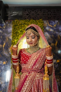 A bride wearing a traditional red and gold embellished lehenga with intricate jewelry, including a heavy necklace and earrings. Her hands are adorned with mehndi and bangles, and she holds a part of her veil with a serene expression. The background features a decorative panel and greenery.